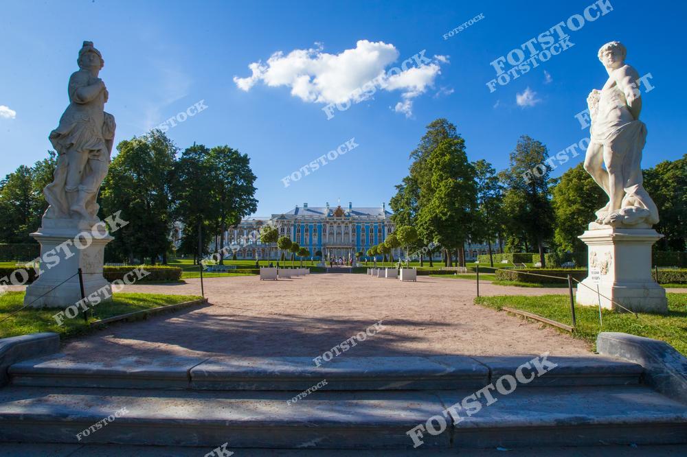 На переднем плане изображены мраморные статуи, обрамляющие вход в регулярную часть Екатерининского парка в городе Пушкине. Вдали виден фасад Екатерининского дворца — знаменитого образца елизаветинского барокко. Пространство между статуями ведёт к дворцу, создавая симметричную композицию. Пышные кроны деревьев подчёркивают монументальность ансамбля. Снимок выполнен летом в ясную солнечную погоду.